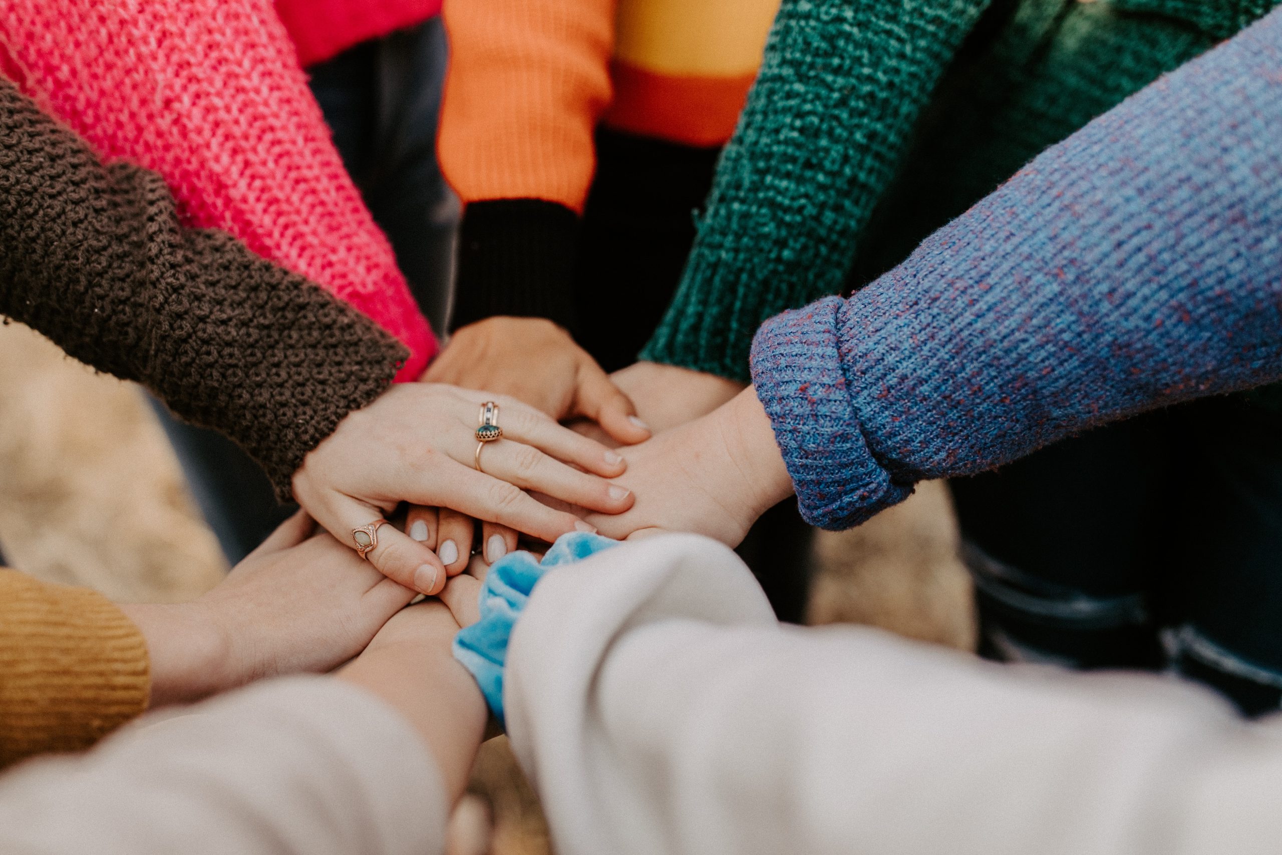 A close-up of several people wearing colorful sweaters, all putting their hands together in a circle for a group gesture.