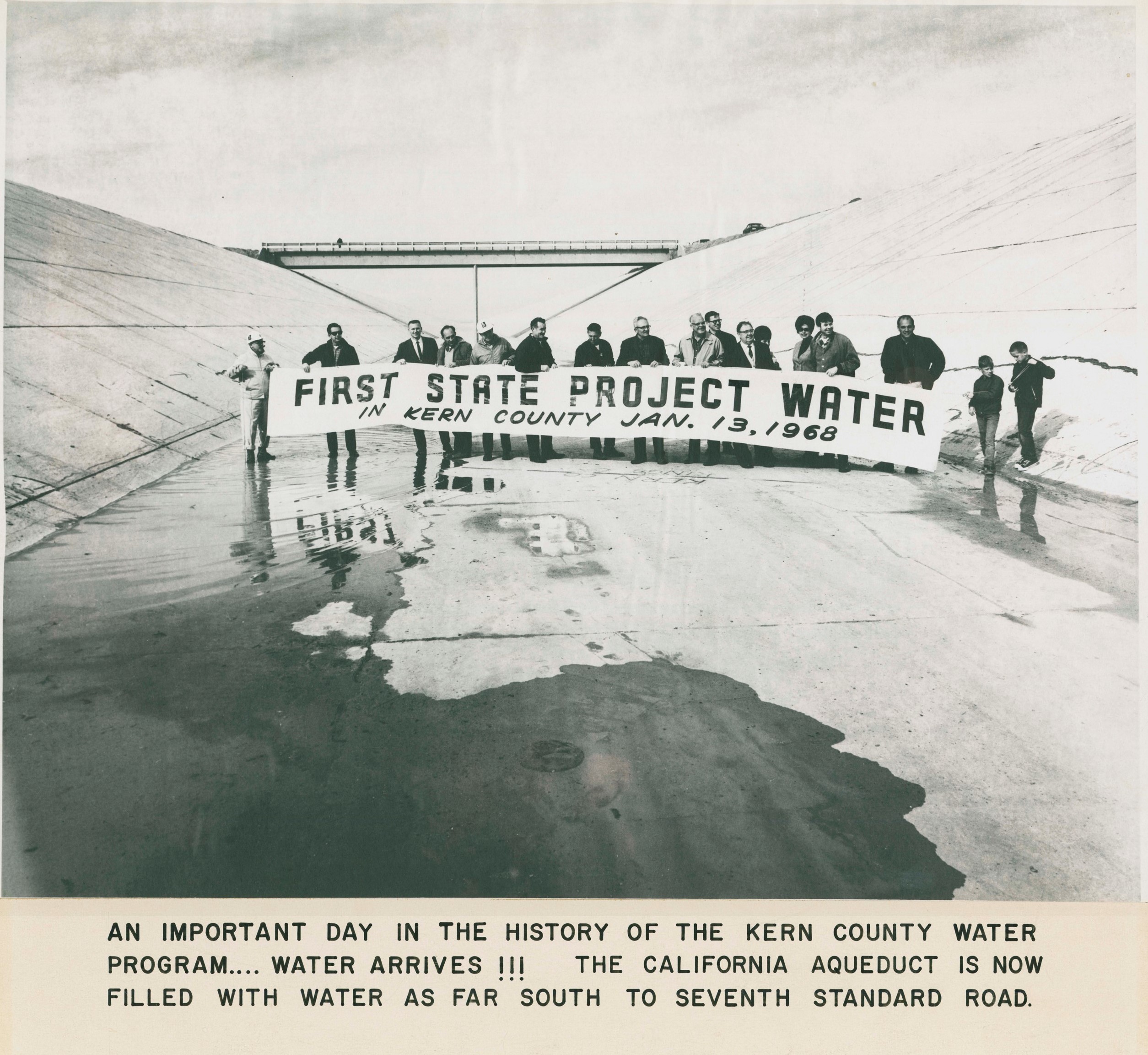 Group of people holding a banner that reads "First State Project Water in Kern County Jan. 13, 1968" in a large canal with water arriving with the words reading "An important day in the history of the Kern County Water Program... water arrives!!! The California Aqueduct is now filled with water as far south to Seventh Standard Road." at the bottom of the image.