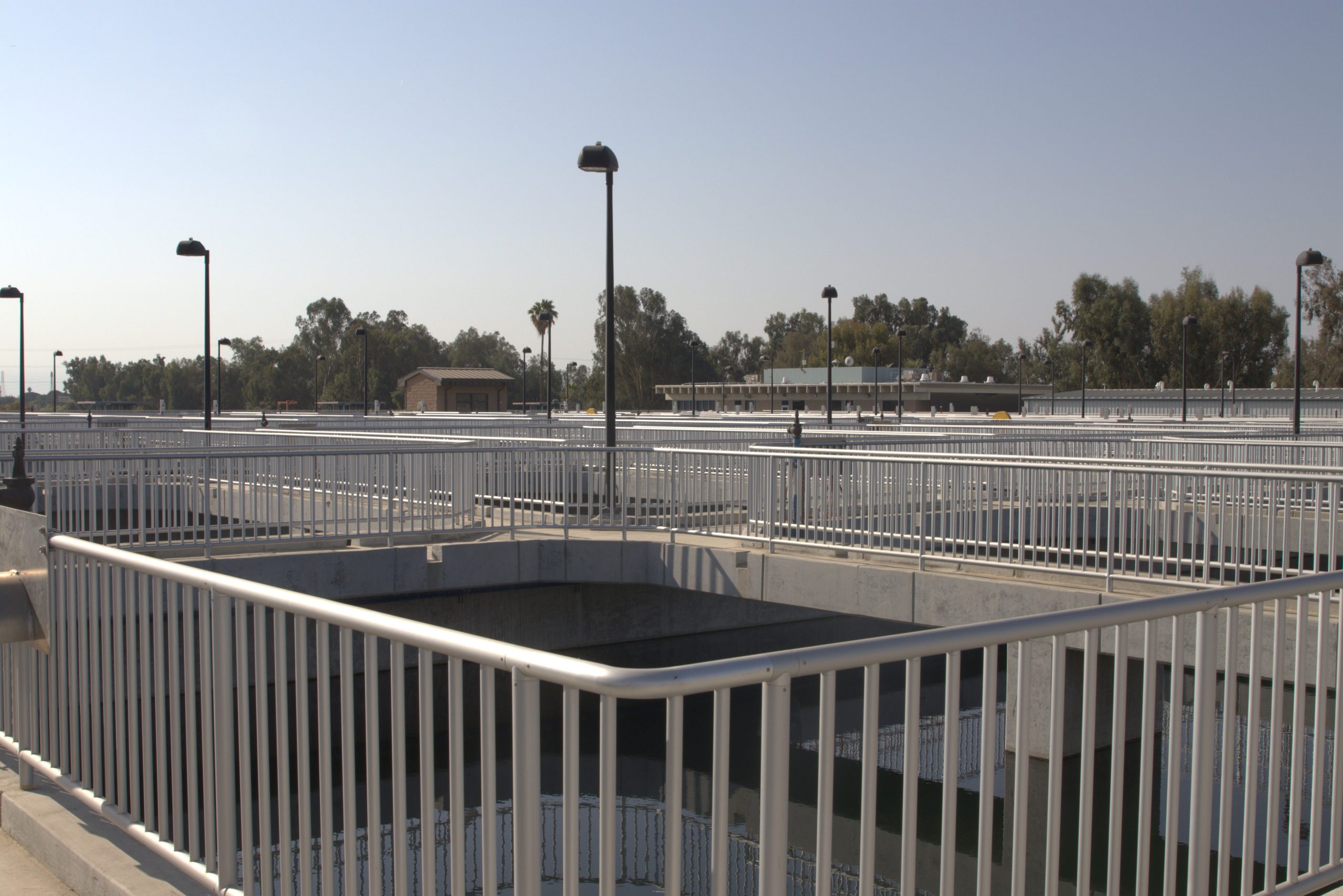 A large water treatment facility with multiple rectangular reservoirs, surrounded by metal railings and lampposts under a clear sky.