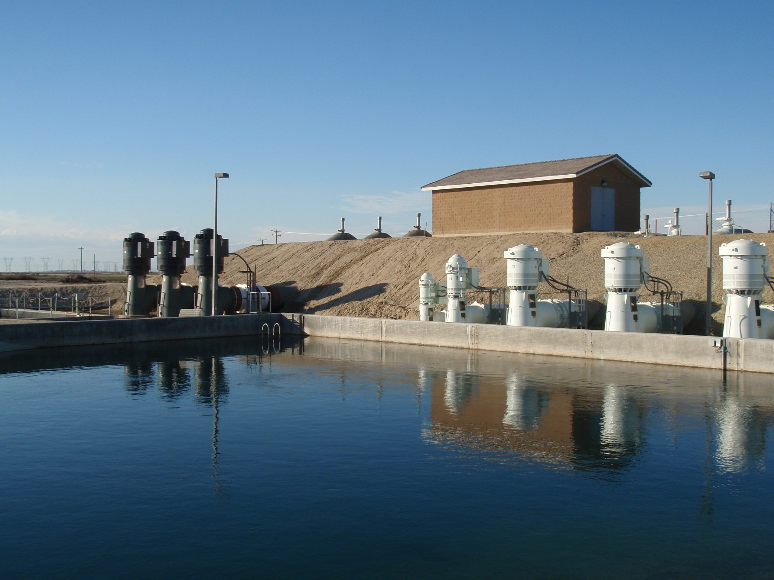 Outdoor water treatment facility featuring a reservoir, several cylindrical tanks, and a small building in the background under a clear blue sky.