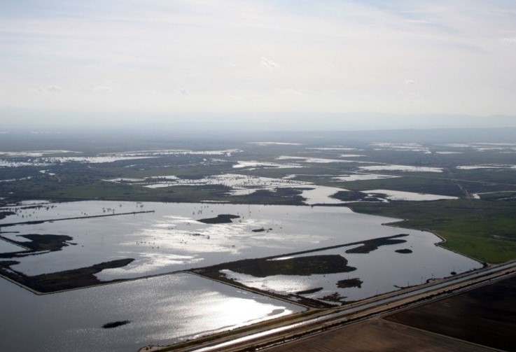 Aerial view of a landscape with flooded fields and water bodies reflecting sunlight on a partly cloudy day.