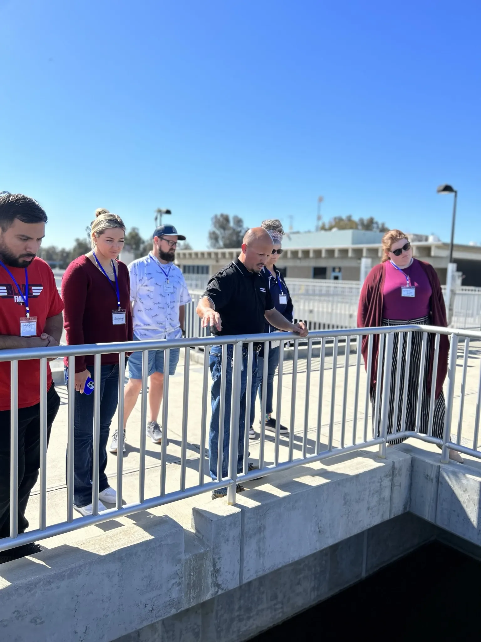 Seven people stand on a walkway with railings, peering over the edge. The sky is clear and blue. They appear to be observing something below, possibly participating in a Project WET activity focused on water education and environmental education.