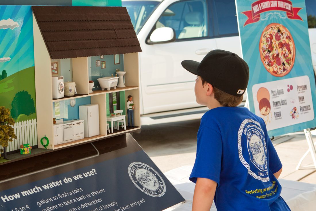 A child in a blue shirt and black cap looks at a detailed cutaway model of a house, displayed at a community exhibit. Informational posters about local events are visible around him.