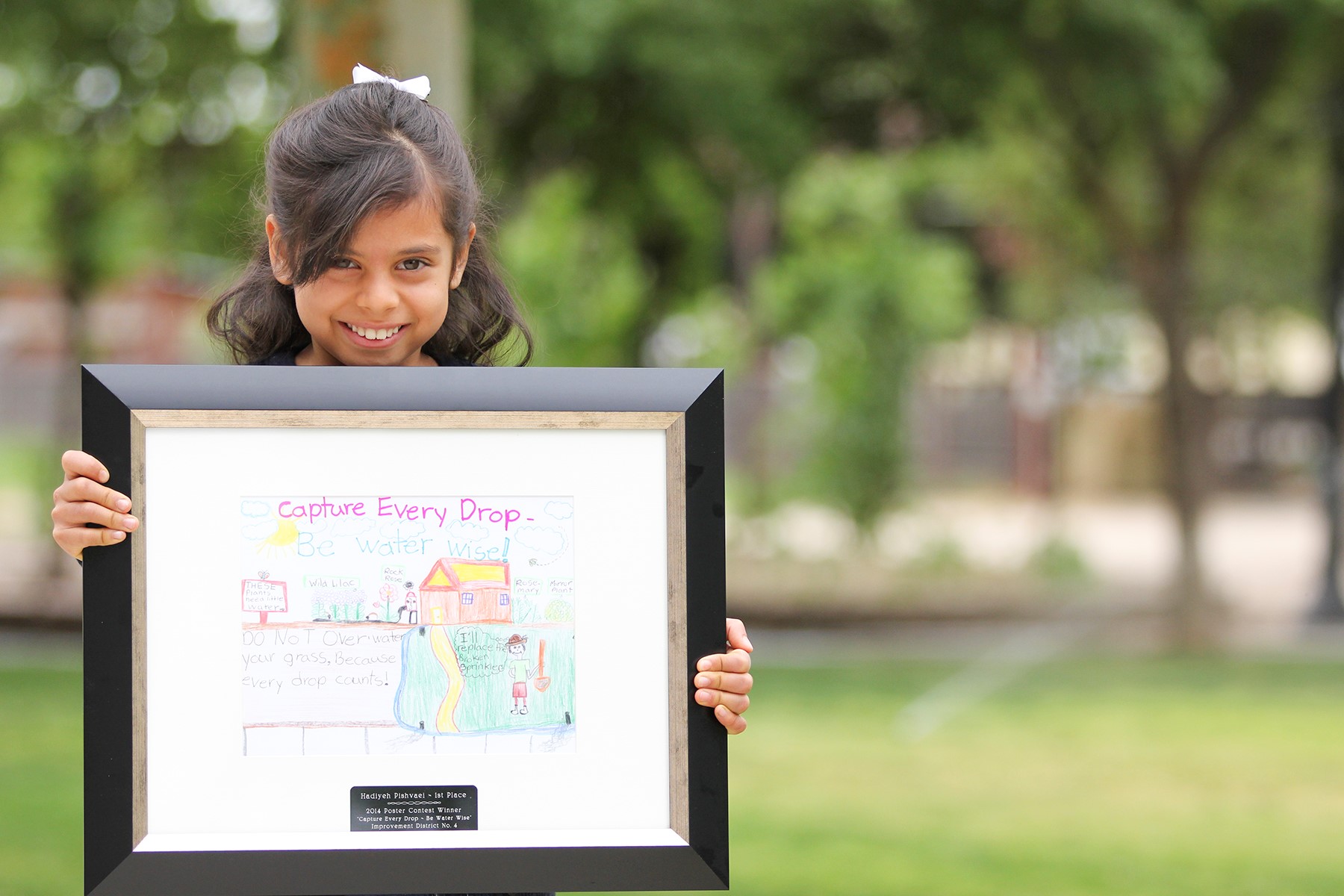 A young girl stands outdoors by her home, holding a framed drawing with a message about water conservation. The drawing features colorful illustrations and text promoting saving water.